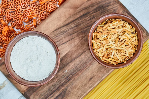 Geassorteerde ongekookte pasta 's en een kom bloem op het houten bord — Stockfoto