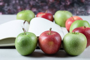 Masada kırmızı ve yeşil elmalar ve boş bir yemek tarifi kitabı.