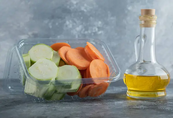 Sliced zucchini and carrot in plastic container and pottle of oil over grey background — Stock Photo, Image