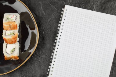 Ejderha suşi tabağı, yılan balığı ve siyah yüzey üzerine boş bir defter ile yuvarlanıyor.