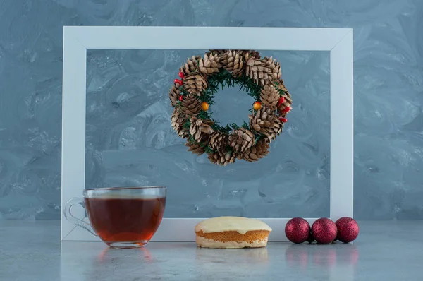 Christmas wreath hanging from an empty frame behind baubles, white chocolate coated cake and a cup of tea marble background. High quality photo