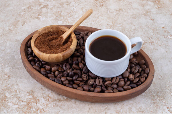 A cup of coffee and a small bowl of ground coffee powder on a pile of coffee beans in a tray on marble background. High quality photo