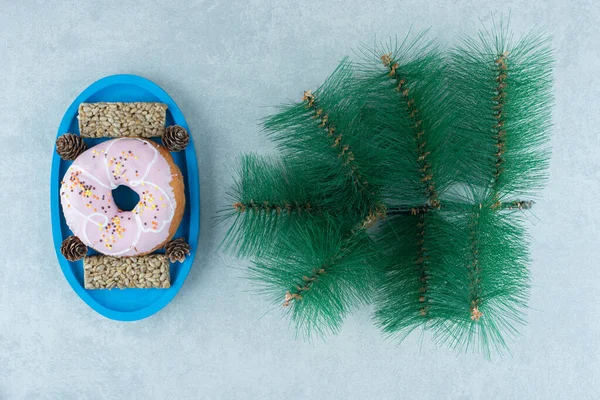 Sunflower seed snack bars and a donut with pine cones on a platter next to an artificial pine branch on marble background. High quality photo
