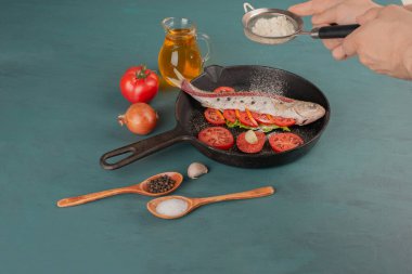 Kadın siyah tavada ızgara balığa un, domates dilimleri, bir bardak yağ ve baharat ekliyor. Yüksek kalite fotoğraf