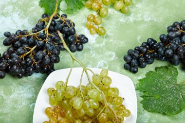 Yeşil arka planda yaprakları olan bir tabak beyaz üzüm ve siyah üzüm.