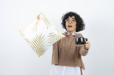 Beyaz arka planda bir fincan kahve ve yastıkla poz veren genç bir kadın.