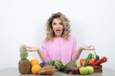 Masada bir sürü meyve ve sebzeyle oturan pembe tişörtlü sarışın bir kadın resmi. Yüksek kalite fotoğraf