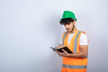 Beyaz duvarın üzerinde deftere yazılı, kasketli yakışıklı erkek mühendis. Yüksek kalite fotoğraf