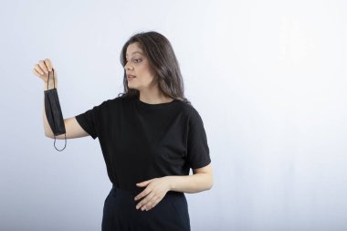 Güzel esmer kız beyaz duvarın üzerinden tıbbi maskeye bakıyor. Yüksek kalite fotoğraf