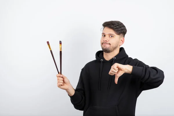 Imagem Jovem Artista Bonito Segurando Pincéis Dando Polegares Para Baixo — Fotografia de Stock