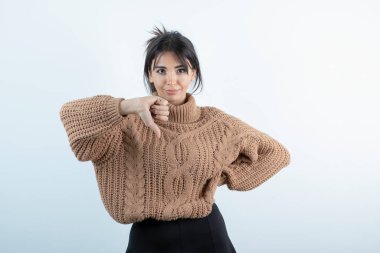 Örgü süveter giyen güzel genç bir kadın beyaz duvarın üzerinden başparmak kaldırıyor. Yüksek kalite fotoğraf