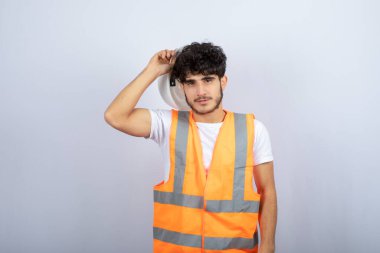 Beyaz kaskı beyaz duvarın üstünde tutan üniformalı yakışıklı bir genç. Yüksek kalite fotoğraf