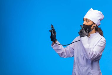 Siyah maskeli ve eldivenli genç hemşire ağzını kapatırken hastayı steteskopla kontrol ediyor. Yüksek kalite fotoğraf