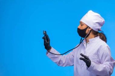 Tecrit edilmiş hemşire, elinde steteskopla ve yüzünde maskeyle hastayı dikkatlice kontrol ediyor. Yüksek kalite fotoğraf