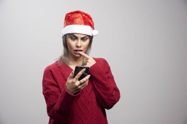 Kırmızı kazaklı genç bir kadın gri arka planda cep telefonuna bakıyor. Yüksek kalite fotoğraf