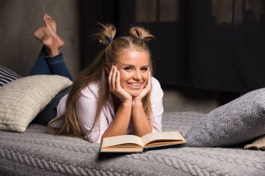 Genç bir kadın, karanlık arka planda bir kitapla yatakta yatıyor.