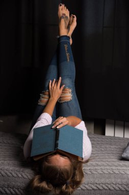 Yatağa uzanmış bir kadın ve karanlık arka planda yüzüne kitap koyuyor.