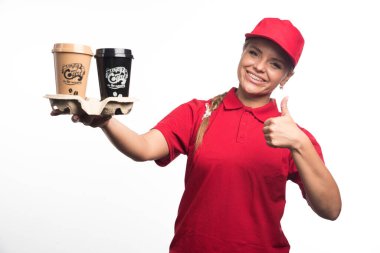 Kadın kurye elinde iki fincan kahve tutuyor ve beyaz arka planda başparmak kaldırıyor. Yüksek kalite fotoğraf