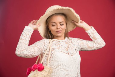Kadın aşağı bakıyor ve kırmızı arka planda şapka ve çantayla poz veriyor.