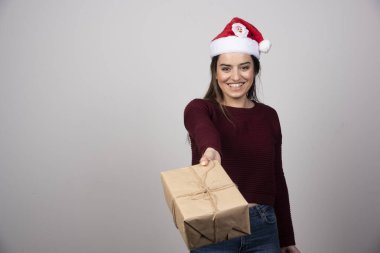Noel Baba şapkalı genç bir kadın gri arka planda Noel hediyesi sunuyor. Yüksek kalite fotoğraf
