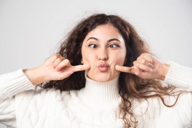 Beyaz kazaklı genç bir kadın beyaz bir duvarın üstünde komik bir surat yapıyor. Yüksek kalite fotoğraf