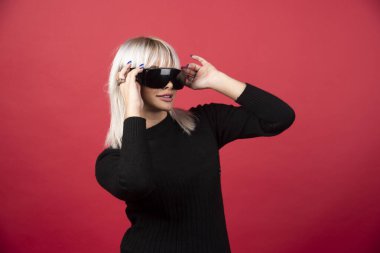 Kırmızı arka planda siyah gözlüklü, ayakta duran ve poz veren kadın portresi. Yüksek kaliteli fotoğraf.