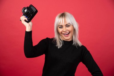  Güzel genç bir kadın kırmızı arka planda dururken elinde bir kamera tutuyor. Yüksek kalite fotoğraf