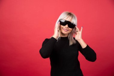 Kırmızı arka planda siyah gözlüklü, ayakta duran ve poz veren kadın portresi. Yüksek kaliteli fotoğraf.