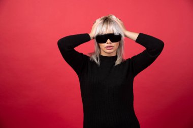 Kırmızı arka planda siyah gözlüklü, ayakta duran ve poz veren kadın portresi. Yüksek kaliteli fotoğraf.