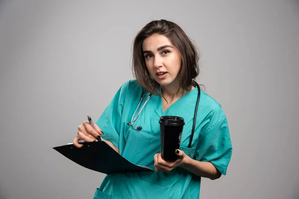 灰色の背景にカップとクリップボードを保持する若い医師 高品質の写真 — ストック写真