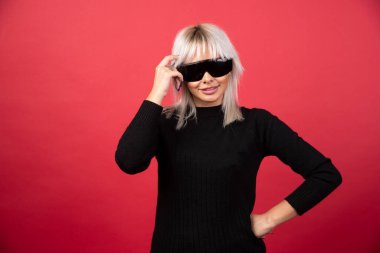 Kırmızı arka planda siyah gözlüklü, ayakta duran ve poz veren kadın portresi. Yüksek kaliteli fotoğraf.