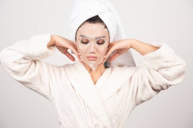 Yüzüne maskeyle dokunurken bornozlu ve kafasında havlu olan genç bayanın portresi. Yüksek kalite fotoğraf