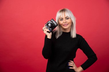  Güzel genç bir kadın kırmızı arka planda dururken elinde bir kamera tutuyor. Yüksek kalite fotoğraf