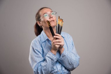 Kadın sanatçı, gri arka planda yüzünün yakınında fırça boyası tutuyor.