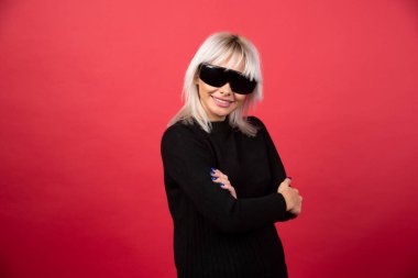 Kırmızı arka planda siyah gözlüklü, ayakta duran ve poz veren kadın portresi. Yüksek kaliteli fotoğraf.