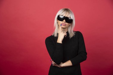 Kırmızı arka planda siyah gözlüklü, ayakta duran ve poz veren kadın portresi. Yüksek kaliteli fotoğraf.