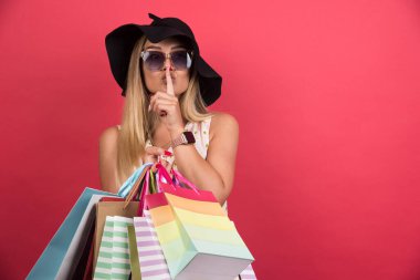 Gözlüklü ve şapkalı bir kadın alışveriş çantalarını tutuyor kırmızı arka planda sessizlik işareti yapıyor. Yüksek kalite fotoğraf