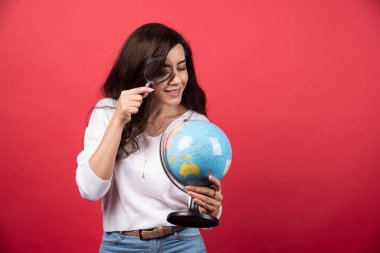 Genç bir kadın model kırmızı arka planda büyüteçle bir küreye bakıyor. Yüksek kalite fotoğraf