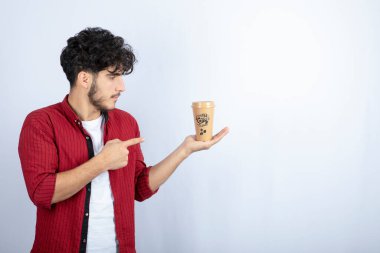 Sıradan kıyafetli genç adamın resmi beyaz arka plandaki bir fincan kahveyi işaret ediyor. Yüksek kalite fotoğraf
