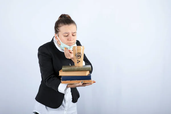 Image of young woman holding books and cup of coffee and taking off mask. High quality photo