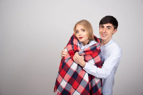 Young man covering freezing girlfriend with plaid blanket while standing against gray wall. High quality photo