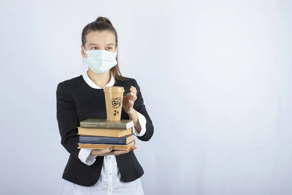 Image of young woman in face mask holding books and cup of coffee over white wall. High quality photo
