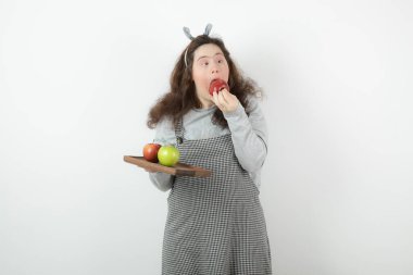 Kıvırcık saçlı, ayakta duran ve elma meyvesi yiyen tatlı bir kızın fotoğrafı. Yüksek kalite fotoğraf