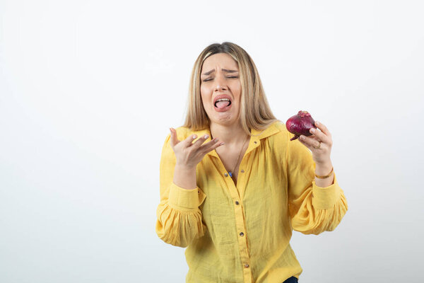 Portrait of blonde woman holding red onion with disgusted expression. High quality photo