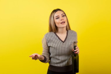 Sarı arka planda şarap şişesiyle poz veren güzel bir kadın.