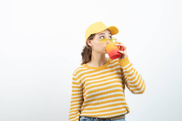 Image of cute young woman in glasses standing and drinking from a mug . High quality photo