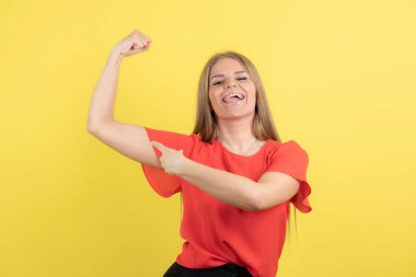 Sarı duvardaki pazılarını gösteren ve gülümseyen genç bir kadının fotoğrafı. Yüksek kalite fotoğraf