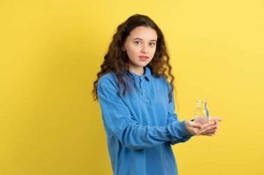 Sarı duvarda mavi kazaklı, kıvırcık bir gencin bir bardak su taşıdığı fotoğraf. Yüksek kalite fotoğraf