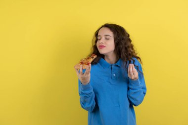Genç kızın portresi bir dilim pizza tutuyor ve sarı arka planda kokusunu alıyor. Yüksek kalite fotoğraf