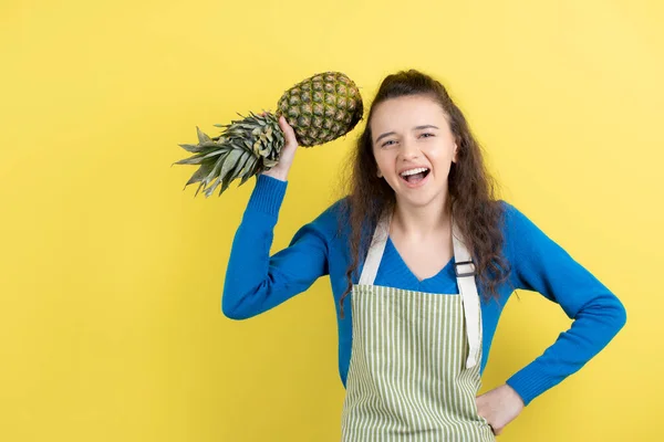 Zdjęcie Młodego Kręconego Nastolatka Fartuchu Trzymającego Dojrzały Ananas Nad Żółtą — Zdjęcie stockowe